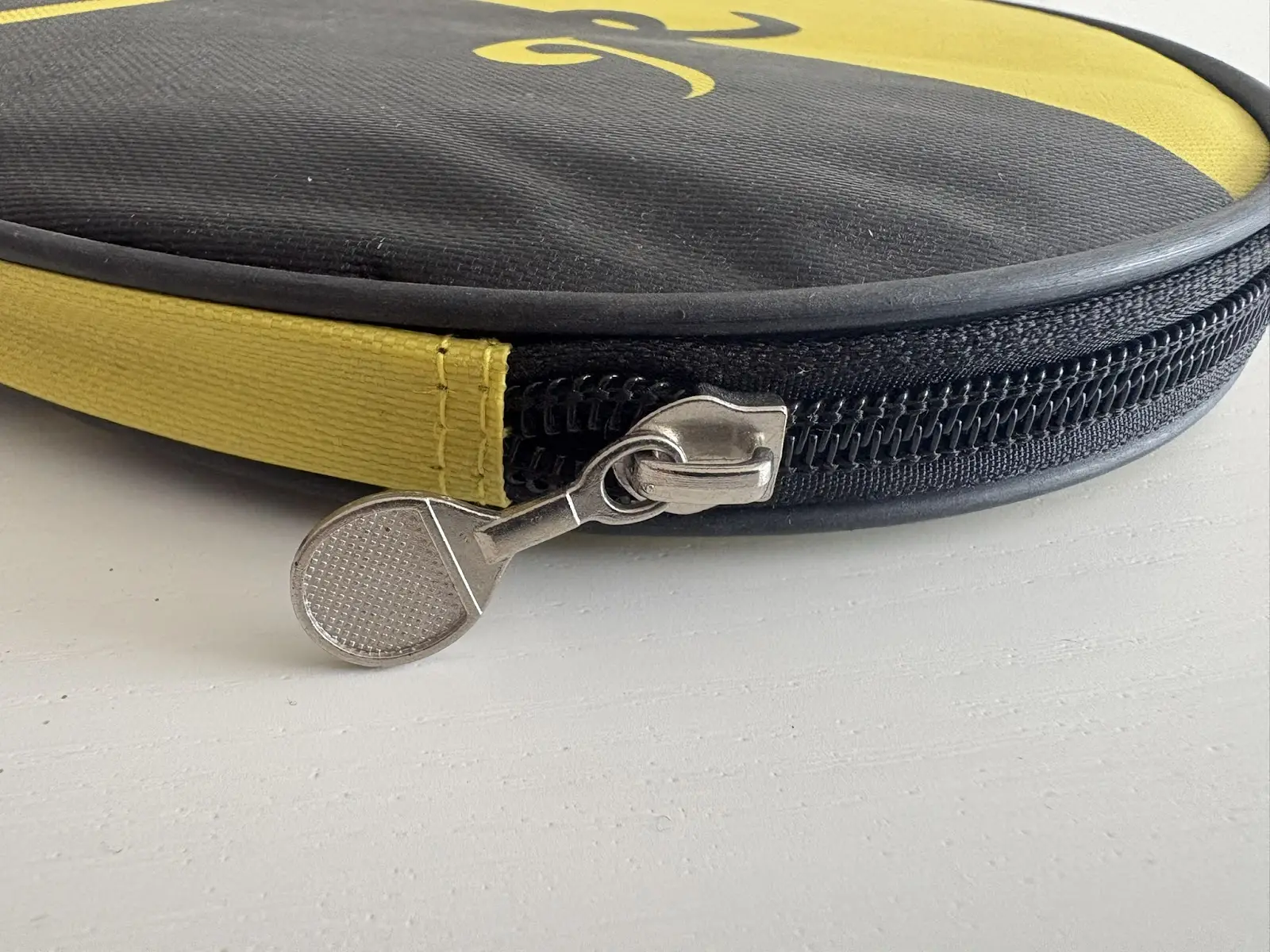 Close-up of a black and yellow round zippered case on a white surface, showing black zipper teeth, stitched yellow fabric edging and a silver metal zipper pull shaped like a small racket.