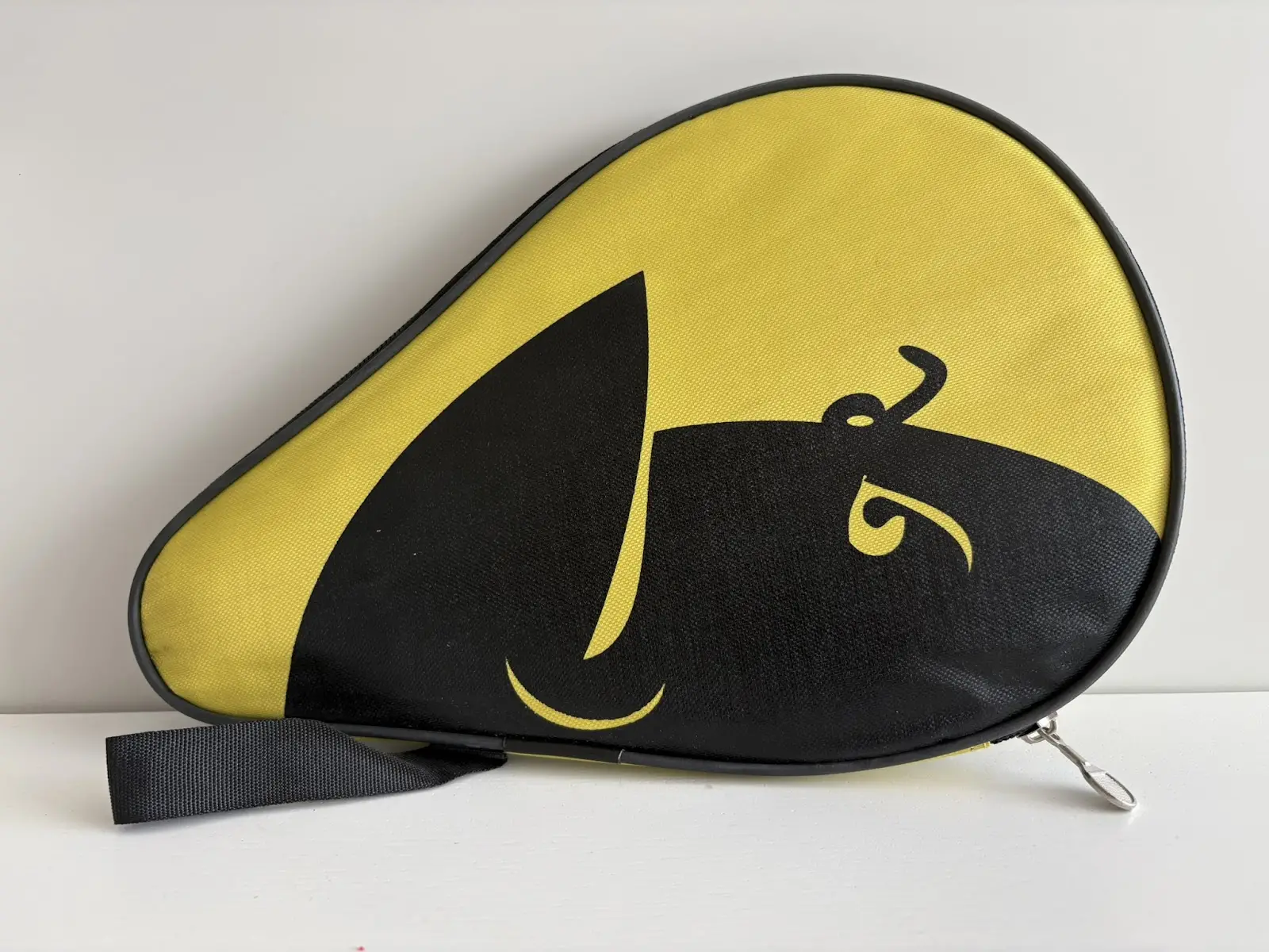 Teardrop-shaped yellow fabric case with a large black abstract graphic, black edging, a short black wrist strap and a metal zipper pull, photographed on a white surface.
