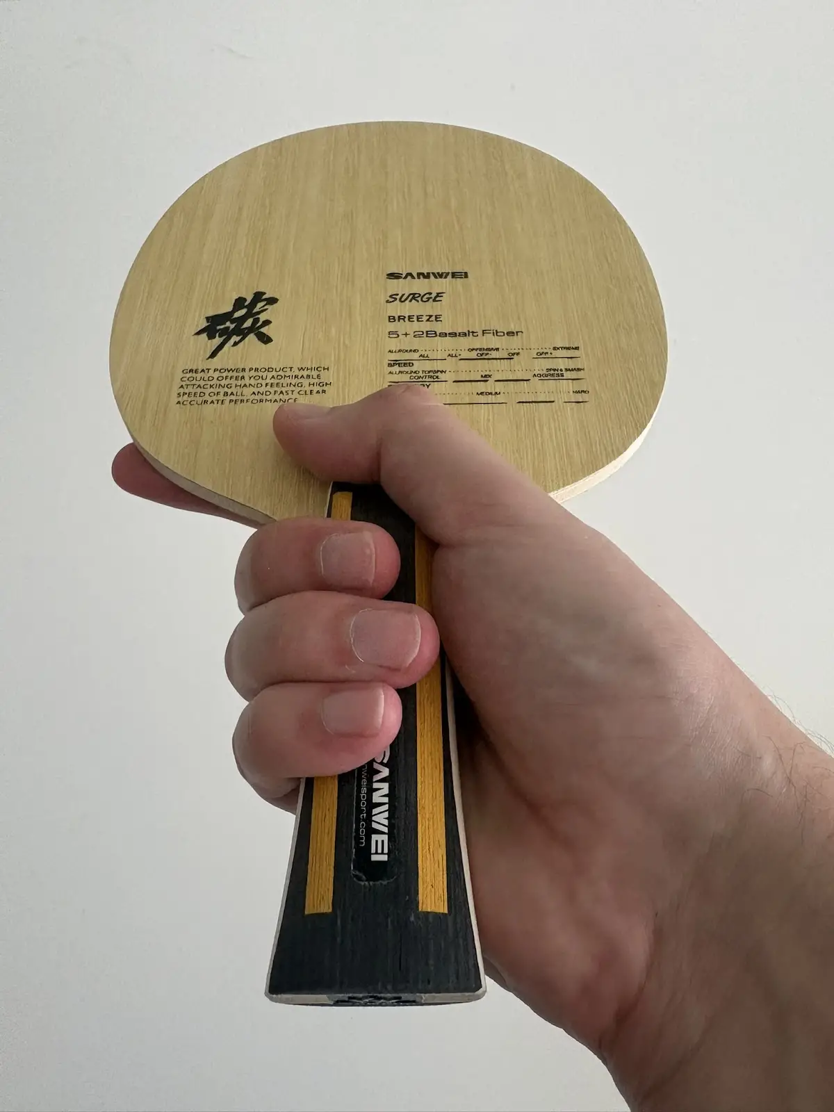 A hand holding a bare wooden Sanwei Surge Breeze table tennis blade by its black-and-yellow handle, showing printed model text and a Chinese character on the paddle face.